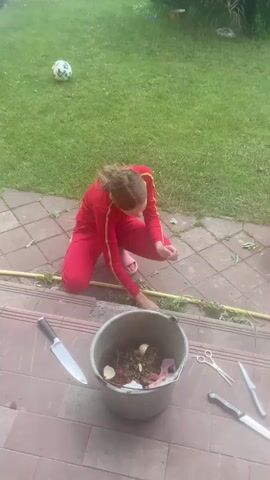The video shows a young person in a red tracksuit kneeling on a paved area in front of a set of outdoor steps. They are looking down and appear to be working with something on the ground. Beside them is a large grey bucket filled with what looks like organic matter, along with some white eggshells. There are also several knives and a pair of scissors laid out on the steps. In the background, a red toy wheelbarrow is visible on the grass, and a soccer ball is on the lawn further back. The overall environment is an outdoor, grassy yard. Later in the video, a second person, a young boy in a blue shirt and green pants, is seen kneeling further away on the paved area, also appearing to be engaged in an activity on the ground.