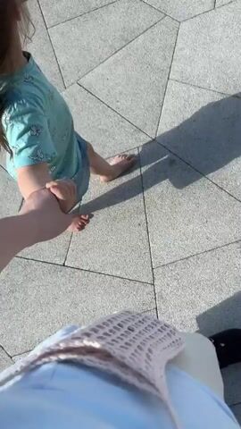 The video shows a child's bare feet walking on a paved walkway. The child is wearing a light blue t-shirt with a pattern and denim shorts. The child's hand is being held by an unseen person, implied to be the person filming. Another person's legs and feet, wearing black sandals, are visible further ahead. Shadows of people are cast on the pavement, indicating a sunny day. The paving stones are grey and arranged in a geometric pattern. The camera angle is low, focusing on the child's feet and the ground.