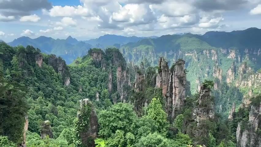 The video shows a scenic view of a mountainous landscape with tall, pillar-like rock formations covered in lush greenery. In the background, there are rolling hills and mountains receding into the distance under a cloudy sky. A woman speaks briefly in Mandarin.