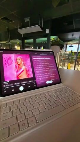 A tablet computer rests on a keyboard, displaying the lyrics to the song "Streets" by Doja Cat. The album art, a pink-toned image of the artist, is visible on the left side of the screen. The tablet is positioned on a table, likely in a public setting, suggested by the presence of what appears to be a McDonald's restaurant in the background. The ambient lighting is dim, with the screen of the tablet being the primary light source in the foreground. The keyboard is white and appears to be a detachable accessory for the tablet.