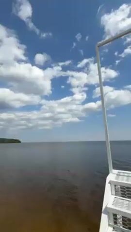 The video shows a wide body of water with a cloudy blue sky above. In the foreground, there are white metal stairs with a square frame extending upwards. The water appears calm, with a slight ripple on the surface. The horizon is visible in the distance. The camera is still.