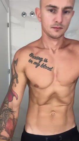 A man with a shaved head and visible abdominal muscles is standing in a room, likely a bathroom, with a white tiled wall behind him. He is shirtless and wearing black shorts. He has tattoos on his chest and left arm. The tattoo on his chest reads "Boxing is in my blood". The man is looking directly at the camera and appears to be flexing his abdominal muscles, causing his rib cage to become more prominent. The video shows a close-up of the man's upper body as he performs this action.