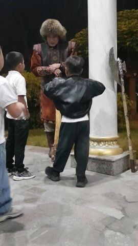 A group of people stand near a large white pillar with gold accents and an axe mounted on top. Two young boys in white t-shirts and jeans are present. One boy is facing forward, while the other is turned away from the camera, appearing to be interacting with someone off-screen. A third person, possibly an adult, is dressed in historical attire, including a fur hat and armor. This person is also turned away from the camera. The ground is paved with irregular stones, and there are trees in the background. The scene appears to be outdoors at night.