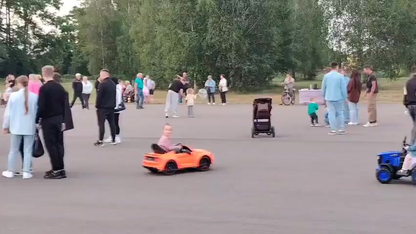 The video shows an outdoor gathering of people in what appears to be a rural setting, possibly a village fair or festival, in Олтушскі сельскі Савет, Belarus. The scene is set in a paved area surrounded by trees, suggesting a park or an open field.

The subjects are a diverse group of people of all ages, including adults, teenagers, and young children. Many are standing and interacting, some in small groups, others in larger clusters. A few children are engaged in recreational activities. One child is seen riding in a small, bright orange toy car, while another is seated in a black stroller. In the background, there are several vehicles, including a white van with a serving window that resembles a food truck, and a white bus. There's also a small stage or platform with a large colorful banner or backdrop, and two tents, one red and one green.

The activity appears to be a community event, with people gathered and seemingly enjoying themselves. The time of day seems to be late afternoon or early evening, as indicated by the soft lighting and the long shadows. The weather appears to be pleasant, conducive to an outdoor event.

Notable details include the presence of a stage area with signage that is partially visible, featuring text that appears to be in the Cyrillic alphabet. While the specific text is not fully legible, it could be related to the event or its sponsors. The overall mood seems relaxed and festive, with people congregating and enjoying the occasion. The setting is distinctly Belarusian, with the language cues and the typical village event atmosphere.