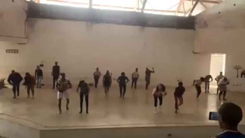 The video depicts a group of around 16 people dancing on a stage-like platform in a large, open indoor space. The space appears to be a hall or gymnasium, with white walls, a corrugated metal roof, and simple decorations near a doorway on the right side of the shot. A person in the foreground is recording the group with a smartphone. The dancers are dressed in casual attire and are performing a coordinated routine. The lighting appears natural, coming from windows or openings in the building. The overall atmosphere seems energetic and positive.