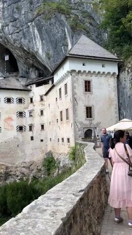 The video opens with a shot of Predjama Castle, a Renaissance castle built within a cave in southwestern Slovenia. The castle is perched dramatically on a cliff face, with its white walls contrasting against the gray rock and lush green trees. The camera pans across the castle's facade, highlighting its numerous windows and the cave opening behind it. A person wearing a backpack is visible walking along a stone pathway in the foreground. The video then shifts to a wider view of the surrounding mountainous landscape, showing dense forests and a clear blue sky with fluffy white clouds. The overall impression is one of natural beauty and historical grandeur.