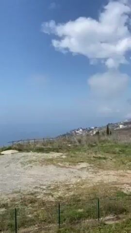 The camera pans slowly across a scenic view of the ocean. The sky is blue with scattered white clouds. A rough, stony surface occupies the foreground, partially enclosed by a low green fence. Beyond this area, a simple wooden fence marks the edge of a grassy area overlooking the ocean. In the distance, a coastline is visible. A tree is visible on the left-hand side.
