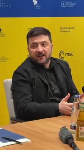 The video shows Volodymyr Zelenskyy, the President of Ukraine, sitting at a table. He is wearing a black shirt and a black jacket. Behind him is a backdrop with various logos, including "Munich Security Conference" and "Office of the President of Ukraine." On the table in front of him are a bottle of water, a bottle of soda, a pen, a blue folder, and a microphone. Zelenskyy is speaking and gesturing with his hands. He then looks to the side, apparently listening. The camera angle is a medium shot.