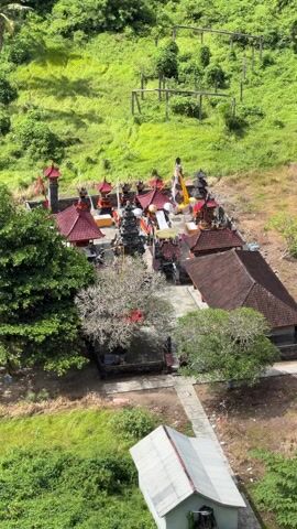 An eye-level aerial shot showcases a Balinese temple complex nestled amidst lush greenery. The temple features multiple structures with red-tiled roofs and decorative details. Tall black stone towers are interspersed among the buildings. People can be seen walking around the temple grounds, and a tall, light-colored structure stands prominently within the complex. The surrounding landscape is characterized by vibrant green grass and trees. A small, white-roofed building sits slightly apart from the main complex. The overall scene depicts a serene and cultural setting.