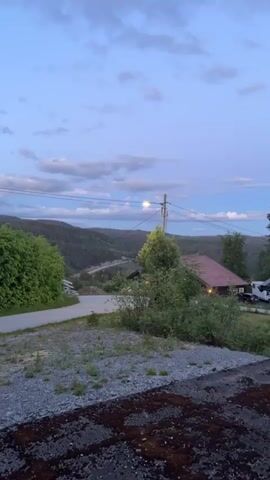The video pans across a rural landscape at dusk. In the foreground, there is a gravelly area with sparse vegetation. A paved road curves to the left. In the mid-ground, there are trees and bushes, a building with a reddish-brown roof, and another smaller dark-colored building to the right. A utility pole with wires and a street light is visible. In the background, a valley is filled with dense coniferous forest, and rolling hills stretch towards the horizon under a sky with scattered clouds, tinged with the blues and purples of twilight.