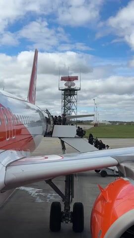 The video shows passengers disembarking an orange and white airplane via a ramp. The plane is parked on the tarmac at an airport. A radar tower is in the background. The sky is blue with puffy white clouds. Wind is blowing and can be heard in the audio. The camera angle shows the side of the plane and the wing.