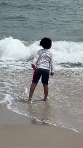 Here is a description of the video:

The video shows a young child standing on a sandy beach, facing the ocean. The child is wearing a white long-sleeved shirt and dark shorts with a red stripe on the side. The waves are crashing onto the shore and the child is getting their feet wet in the surf. The child squats down, touching the water. The sky is overcast and the ocean appears choppy.