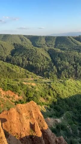 The camera pans left, revealing a vast landscape under a bright, clear sky. In the foreground, dry, reddish-brown earth and rocks are visible, with patches of green vegetation and yellow wildflowers on the upper right. Below, a deep valley is filled with a dense forest of green trees and bushes. In the distance, layers of green hills and mountains stretch towards the horizon, where the sky meets the land.