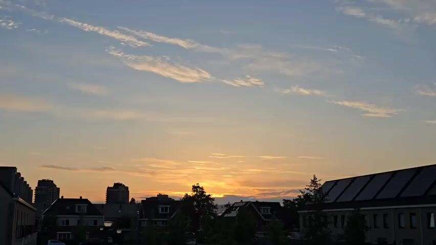 The video shows a suburban or urban landscape at sunset. The sky is mostly a light blue fading into shades of orange and yellow near the horizon where the sun is setting. Wispy clouds stretch across the sky. The silhouettes of buildings, including houses with pointed roofs and taller structures, form the horizon line. The roofs of some buildings are visible. Some trees are also silhouetted. The light is soft. The overall impression is peaceful and serene.