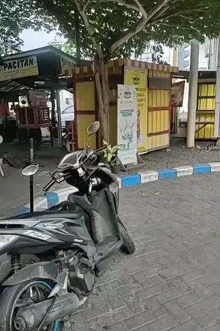 THE VIDEO SHOWS A MOTORCYCLE PARKED ON A PAVED AREA WITH BRICK-LIKE STONES. TO THE LEFT, THERE IS A RED STRUCTURE WITH SIGNS, ONE IN INDONESIAN THAT APPEARS TO BE RELATED TO MOTORCYCLE SERVICES LIKE OIL CHANGES AND NITROGEN FILLING. A MAN WEARING A RED UNIFORM AND A GREEN HAT IS STANDING NEAR THE RED STRUCTURE, FACING AWAY FROM THE CAMERA. FURTHER BACK, THERE ARE SMALL FOOD STALLS OR SHOPS. THE MOTORCYCLE IN THE FOREGROUND IS A DARK COLOR AND HAS A LICENSE PLATE. A HOSE IS CONNECTED TO THE MOTORCYCLE'S REAR WHEEL. TREES AND OTHER BUILDINGS ARE VISIBLE IN THE BACKGROUND. THE GROUND IS A MIX OF PAVERS AND BRICKS. THERE IS ALSO A BLUE AND WHITE BORDER AT THE EDGE OF THE PAVED AREA.