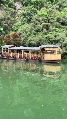 The video shows a tranquil scene of a river or lake surrounded by lush greenery and steep cliffs. The water is a distinctive green hue. A long, covered boat is docked alongside a small wooden structure on the shore. The boat has a series of life preservers attached to its exterior. The camera pans slightly to the left, revealing more of the shoreline, the wooden structure, and the imposing cliff face. The overall impression is one of calm and natural beauty.