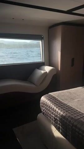 The video shows a luxurious bedroom on a yacht. The room is decorated in neutral tones with dark wood accents. A large bed with quilted bedding dominates the right side of the frame. To the left of the bed, there is a window overlooking the ocean, with a chaise lounge positioned beneath it. A closet is visible on the back wall, and a small desk with a chair is located near the window. The overall impression is one of modern elegance and comfort, with the natural beauty of the sea providing a stunning backdrop.