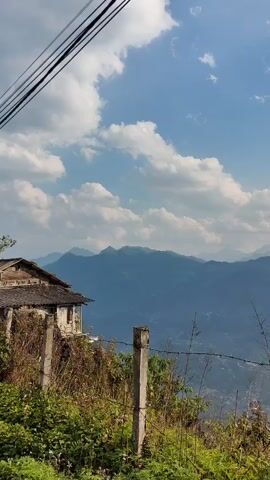 The video shows a mountain landscape with a partly cloudy sky. The sun is shining through the clouds, creating a bright and hazy effect. In the foreground, there's a fence and some overgrown vegetation. Behind the fence, there's an old, slightly dilapidated house. In the background, there's a range of mountains fading into the distance due to atmospheric haze. Above, power lines are strung across the sky. The camera pans upwards during the video, adjusting the view.