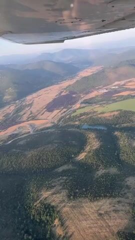The video shows an aerial view from an airplane of a valley with mountains in the background. A highway cuts through the valley, and there are fields and forests visible. The sun is setting, casting long shadows across the landscape.