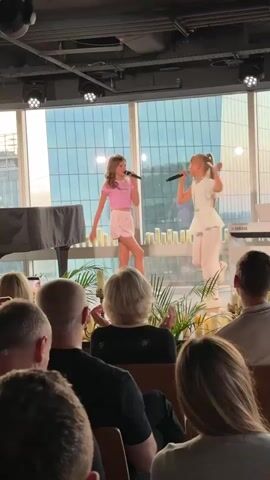 Two young girls are performing on a stage in front of an audience. The girl on the left wears a pink crop top and pink shorts. The girl on the right wears a white ruffled top and white pants. They are both singing into microphones and dancing. Behind them is a large window with a city view, a piano, and a few decorative candles and plants. In front of the stage, the backs of audience members' heads are visible. The lighting appears to be dim, with stage lights focused on the performers.