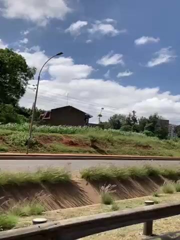 The video shows a view from a moving vehicle of a road with two people walking on the side. The sky is blue with scattered white clouds. Green grass and foliage cover the ground on the side of the road, with a building visible in the distance. There are power lines and street lamps along the road. The vehicle's interior is visible at the bottom of the frame.
