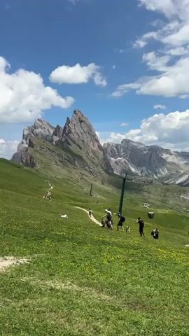 This is a landscape video captured in the Dolomites, specifically Santa Cristina Gherdëina, Italy. The scene is set on a vibrant green mountainside, dotted with yellow wildflowers, under a bright blue sky with scattered white clouds. Dominating the background are majestic, rugged mountain peaks with rocky outcrops and some patches of snow. A ski lift tower is visible on the left side of the frame, suggesting a popular tourist area accessible by lift.

Several people are scattered across the grassy slope, engaged in various activities. Some are walking along a dirt path, while others are seen resting or strolling leisurely on the grass. A group of individuals are gathered in the middle ground, seemingly enjoying the scenic view. In the distance, a rustic building, likely a mountain hut or restaurant, can be observed.

The overall mood is one of a pleasant summer day in a breathtaking natural environment, with people enjoying outdoor recreation. The bright sunlight and blue sky indicate fair weather, likely mid-morning or early afternoon. The presence of a ski lift hints at a location that is active year-round, but the lush green slopes and casual attire of the people suggest a warmer season. There is no visible text in the video.