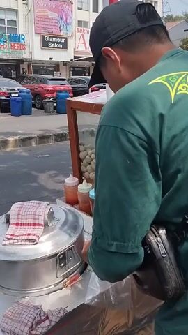 The video shows a street scene. In the foreground, a vendor is preparing food at a street cart. The cart has a metal steaming pot, a display case with food items, and bottles of condiments. A person wearing a green shirt and a black cap is working at the cart. In the background, there are several buildings with shops and businesses. Cars and motorcycles are visible on the street. The environment appears to be an urban area with moderate foot and vehicle traffic. One shot focuses on a woman standing with her arms crossed, looking towards the street. Another shot pans across a row of buildings and shows vehicles moving along the road. The overall impression is of a busy commercial street.