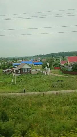 The video shows a landscape view from a moving vehicle, likely a train, looking out over a rural village. The village consists of several houses of various sizes and designs, some with brown roofs and others with blue roofs. The houses are surrounded by green fields, trees, and vegetation. There are also some utility poles visible in the landscape. In the foreground, there is a dirt path or road with some greenery and tall grass. A person is walking on the path in the middle of the frame. The sky is overcast and the overall lighting suggests a cloudy day. The video appears to be filmed horizontally.