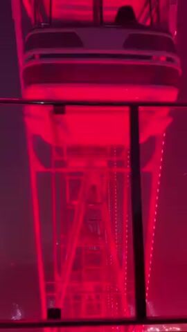 The video shows the view looking down from a moving gondola on a Ferris wheel. The Ferris wheel is lit up in red light, and a safety railing is visible at the bottom of the frame. The structure of the Ferris wheel is visible through the red light. There is a slight fog or haze in the air.