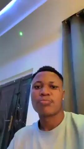 A light-skinned man with short, dark hair is the primary subject of the video. He appears to be inside, possibly a room, as suggested by the visible ceiling with modern blue and white lighting fixtures and a partially visible dark door. He is wearing a light-colored T-shirt and has a stud earring in his left ear. The man is speaking and appears to be singing along to music. The camera angle is low, looking up at the man. There are also dark curtains visible in the background.