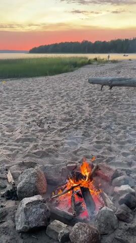 The video shows a campfire burning on a sandy beach at sunset. The sky is a vibrant orange and pink, with a line of trees in the distance. The campfire is made of logs and stones, and there is smoke rising from the flames. The beach is covered in sand, with some footprints visible. There is a fallen log in the background, and a person can be seen standing near the water.