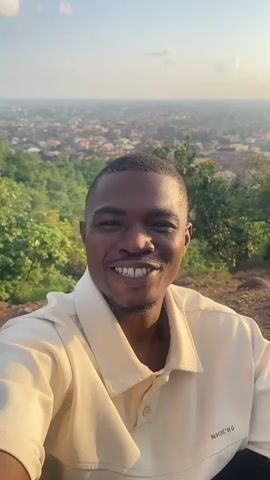 A man wearing a light-colored collared shirt smiles at the camera. He is positioned in the foreground of the video. Behind him, a vast cityscape stretches out into the distance. The cityscape is densely populated with buildings and appears to be nestled within a valley or basin. Trees and rocky terrain are visible on the hillside where the man is standing. The sky is bright, with the sun low in the frame, suggesting it is either early morning or late afternoon. The man turns to wave his hand and gesture towards the city, then turns away from the camera to look out over the landscape. The video appears to be a selfie video, likely taken from a high vantage point overlooking a city.