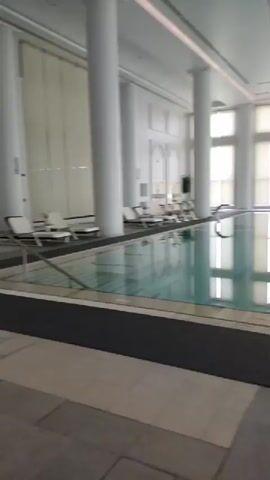 The video shows an indoor swimming pool area. The pool is rectangular and filled with clear blue water, reflecting the light from the large windows and ceiling. The windows are covered with vertical blinds that are partially drawn, allowing diffused light to enter the space. The area around the pool is tiled with a combination of light and dark gray tiles. There are several lounge chairs set up near the pool, some with towels on them, and a few exercise bikes are positioned further away. Large white columns support the ceiling, which has a skylight. The overall atmosphere appears calm and serene, likely a part of a hotel or spa facility.