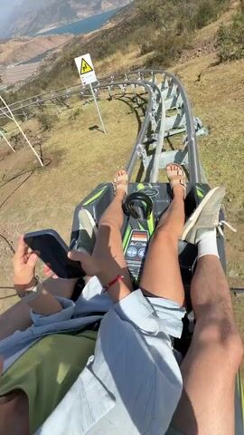 A first-person perspective video shows two people riding a roller coaster. The camera is mounted on the front of the roller coaster cart, facing forward and slightly down. The cart is on a track that winds through a mountainous, dry, grassy landscape. The sky is clear and blue with a few distant mountains. In the foreground, the legs and feet of the two riders are visible. The person in the passenger seat is holding a smartphone and appears to be recording the ride. The cart is moving at a moderate speed, and the surrounding scenery is blurred. The video captures the excitement and thrill of the roller coaster ride.