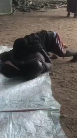 A video shows two children playing on a tarp on a dirt ground. One child, wearing a black shirt and red shorts, is on top of the other child, who is shirtless and lying on their back. The child on top appears to be playing with the other child's hands. In the background, there is a white speaker and other people standing around. There are also piles of debris and tree roots visible in the background.