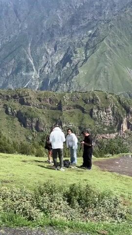 A group of four people stand on a grassy hill. The person on the far left is wearing a dark shirt and white pants. The person next to them is wearing a white hoodie and dark pants. The person in the middle is wearing a white shirt and light-colored pants. The person on the far right is wearing a black shirt and black pants. The group is facing away from the camera and looking at something on the ground, possibly a picnic or a setup for an activity. They appear to be interacting with each other. In the background, there are steep, green mountains with rocky outcrops and patches of snow near the top. The sky is blue with scattered white clouds. The environment is mountainous and natural.