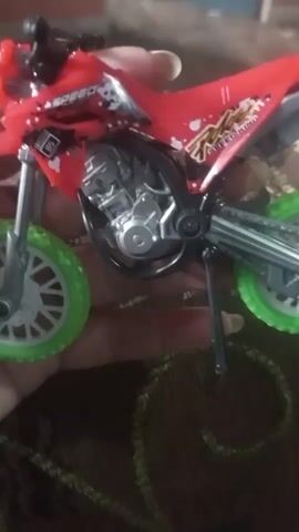 The video shows a close-up of a red toy motorcycle with green wheels. The motorcycle has realistic detailing, including a visible engine and suspension. It appears to be held by a person's hand, as a finger is visible near the handlebars. The background is blurred but seems to be a brown, patterned surface. The narrator expresses delight in the toy, calling it "lucu banget" (very cute) and "keren banget" (very cool).