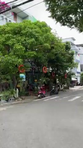 The video shows a street scene, likely in a Vietnamese town or city. The camera is moving, possibly from a vehicle. The focus is on the buildings lining the street and the decorations in front of them. The decorations include plants, red flags, and nautical-themed items like life preservers. There are parked motorcycles on the side of the road. The buildings are a mix of residential and commercial, with some having closed shutters or metal gates. Graffiti is visible on some of the walls. There are also palm trees and utility poles along the street.