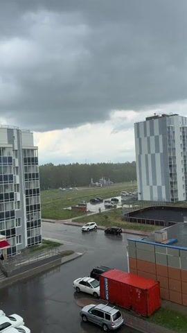 A cloudy sky hangs over a scene of wet streets and buildings. Cars are parked and driving on the wet asphalt. A large red shipping container sits next to a building. In the distance, a forest is visible, and a playground with rocket-shaped structures stands against the trees. The rain is visible as small white specks falling from the sky.