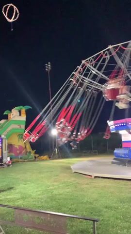 A swing ride at a fairground at night. People are seated on swings that are attached to a central rotating structure. The swings are moving in a circular motion. The ride is lit up with colorful lights. In the background, there is a bouncy castle and some trees. The ground is covered in grass.