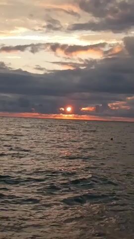 The video shows a sunset over a body of water. The sun is partially obscured by clouds, casting a warm glow on the water's surface. Small waves are lapping at a pebble beach in the foreground. There appears to be a person or object in the water in the distance. The overall scene is serene and picturesque.