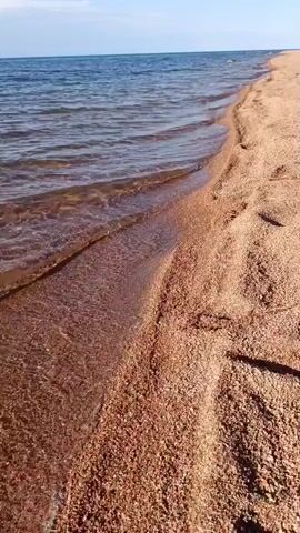 The video shows a close-up view of a sandy beach with gentle waves lapping at the shore. The water is clear and a deep blue, reflecting the sky above. The sand is a warm, reddish-brown color and appears to be made up of small pebbles and grains. Footprints are visible in the sand, suggesting that someone has recently walked along the beach. The sun is shining brightly, casting a warm glow over the scene.