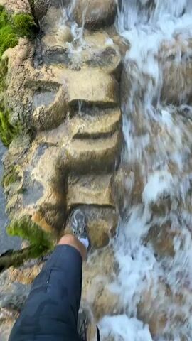 The video shows a first-person perspective of a person walking down a rocky, waterfall-like structure. The person is wearing dark pants and sneakers, one of which is partially visible in each frame as they step down. The environment appears to be natural, with moss growing on the rocks and water flowing down the formation. The camera angle is angled downwards, giving a sense of movement and descent. The lighting is natural, suggesting an outdoor environment. The video is short, only a few seconds long, and captures the act of carefully navigating the terrain.