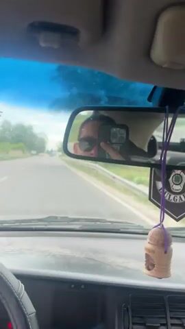 From the interior of a car, a view through the windshield shows a highway under a partly cloudy sky. The car is moving forward, as indicated by the speedometer needle slightly above 60 km/h. There are other vehicles in the distance on the road ahead. The dashboard is visible, and it appears worn and somewhat dusty. The steering wheel is on the left-hand side, suggesting the vehicle is in a region where they drive on the right. Trees and greenery line the sides of the road, and there is a metal guardrail on the right.
