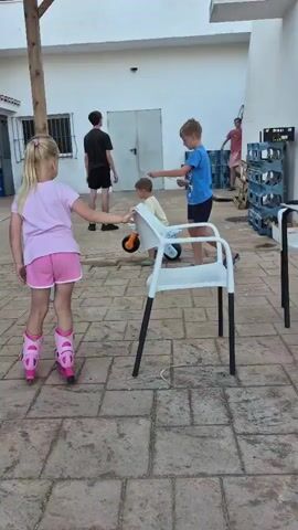 The video shows an outdoor courtyard paved with interlocking stones. Several children are present. In the foreground on the left, a young girl with blonde hair, wearing a pink t-shirt and pink shorts, is on roller skates. She is pushing a white plastic chair with her hands. Beside her is a wooden pole. In the middle ground, a boy in a blue t-shirt and dark shorts is running towards the right. To his left and slightly behind him, another boy in a light-colored shirt and dark shorts is on a small ride-on toy, possibly a scooter or a mini-bike. Further in the background on the right, another person wearing a red shirt and shorts appears to be kicking a ball. To the left of that person, more blue plastic crates are stacked. A large yucca plant with long green leaves is visible on the right side of the frame. The background includes white buildings. The overall scene appears to be a playful or recreational gathering.
