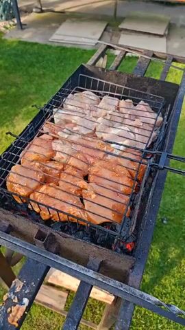 A close-up, high-angle shot shows a metal grill basket filled with chicken pieces cooking over glowing coals. The chicken is arranged in two distinct sections: the front half is marinated in a reddish-brown sauce and appears to be browning, while the back half is lighter in color and appears to be raw or lightly seasoned. The grill is placed on a black metal barbecue stand outdoors, with green grass and a blurred background of outdoor furniture and possibly a patio visible. The scene suggests a warm, sunny day and the preparation of a meal.