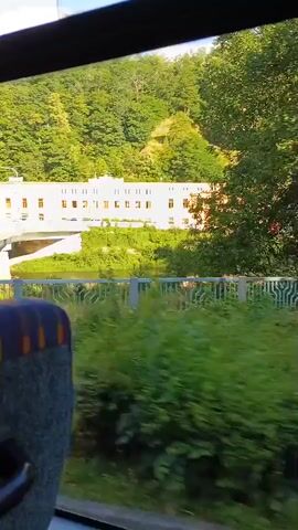 The video shows a view from inside a vehicle, possibly a bus, looking out the window at a scenic landscape. A yellow building with many windows is visible across a body of water. The building is situated on a hillside covered in trees. A bridge is also visible, with a metal railing. The foreground shows the interior of the vehicle, including the back of a seat and part of a window frame. The camera appears to be moving, indicated by the slight blur of the scenery outside. The sky is blue with some white clouds. The overall impression is of a journey through a natural environment with some man-made structures.