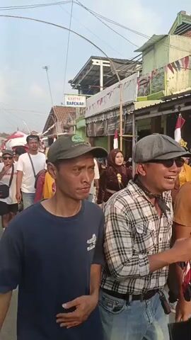 The video shows a street scene with a procession of people. In the foreground, a man wearing a dark blue t-shirt and a green baseball cap is looking to the right. Behind him, a group of people are walking, some of whom are dressed in traditional attire with ornate headpieces and colorful clothing. A man in a white t-shirt and jeans is walking with his arm around another man's shoulder. There are buildings on either side of the street, some adorned with flags. The overall atmosphere appears to be festive or ceremonial.