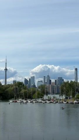 A wide shot captures a picturesque waterfront scene. In the foreground, a tranquil body of water reflects the sky and surrounding elements. A row of sailboats is moored along the shore, their masts standing tall against the backdrop. Beyond the sailboats, lush green trees form a dense line, partially obscuring the impressive Toronto skyline. The iconic CN Tower stands prominently to the left of the frame, piercing the sky. Various skyscrapers, characteristic of a major city, cluster together in the center. The sky above is a mix of blue with scattered white clouds, suggesting a pleasant day. Two individuals are visible in the water, possibly in a small boat or kayak, enjoying the scenery.