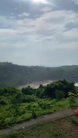 The video shows a scenic camping spot overlooking a beach and ocean. Several tents, primarily red, are set up on a grassy area along a paved path. The camera pans across the scene, revealing the ocean with rock formations in the distance, partially obscured by a light mist. Green foliage covers the landscape between the campsite and the beach. People are visible near the tents, with one person bending down next to a tent. The sky is a pale blue with scattered clouds, and the sun is visible, creating a bright and airy atmosphere.